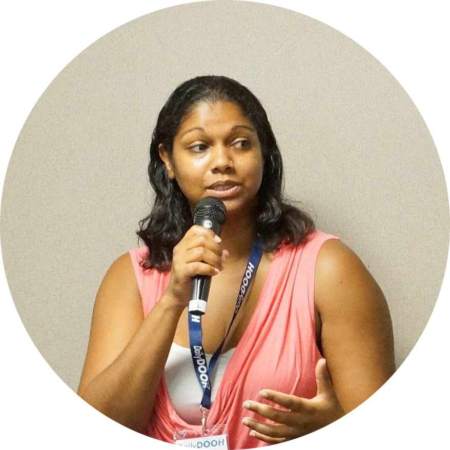 Photo of Helen Hosein, a brown femme with shoulder length hair, speaking into a microphone and wearing a peach top.