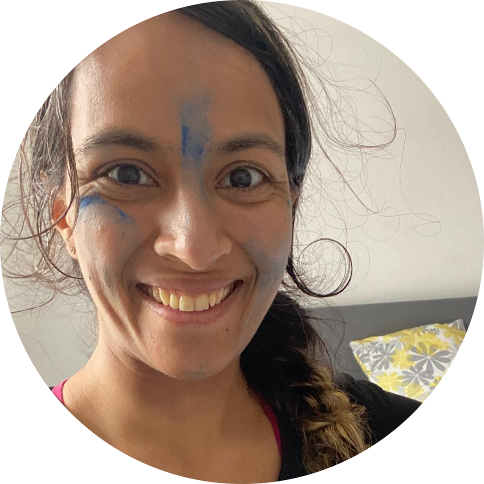 Photo of Alefiyah Jafferji, a smiling brown woman with a ponytail hanging over one shoulder and light smears of blue paint on her face.